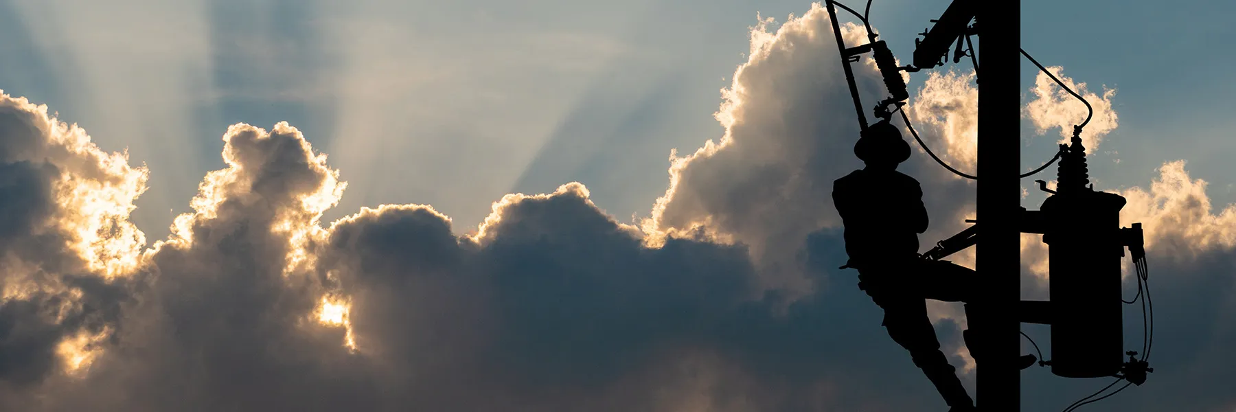 Silueta e një punëtori në shtyllë, me re të fryra dhe rreze dielli në sfond, duke simbolizuar infrastrukturën dhe lidhjen dixhitale.