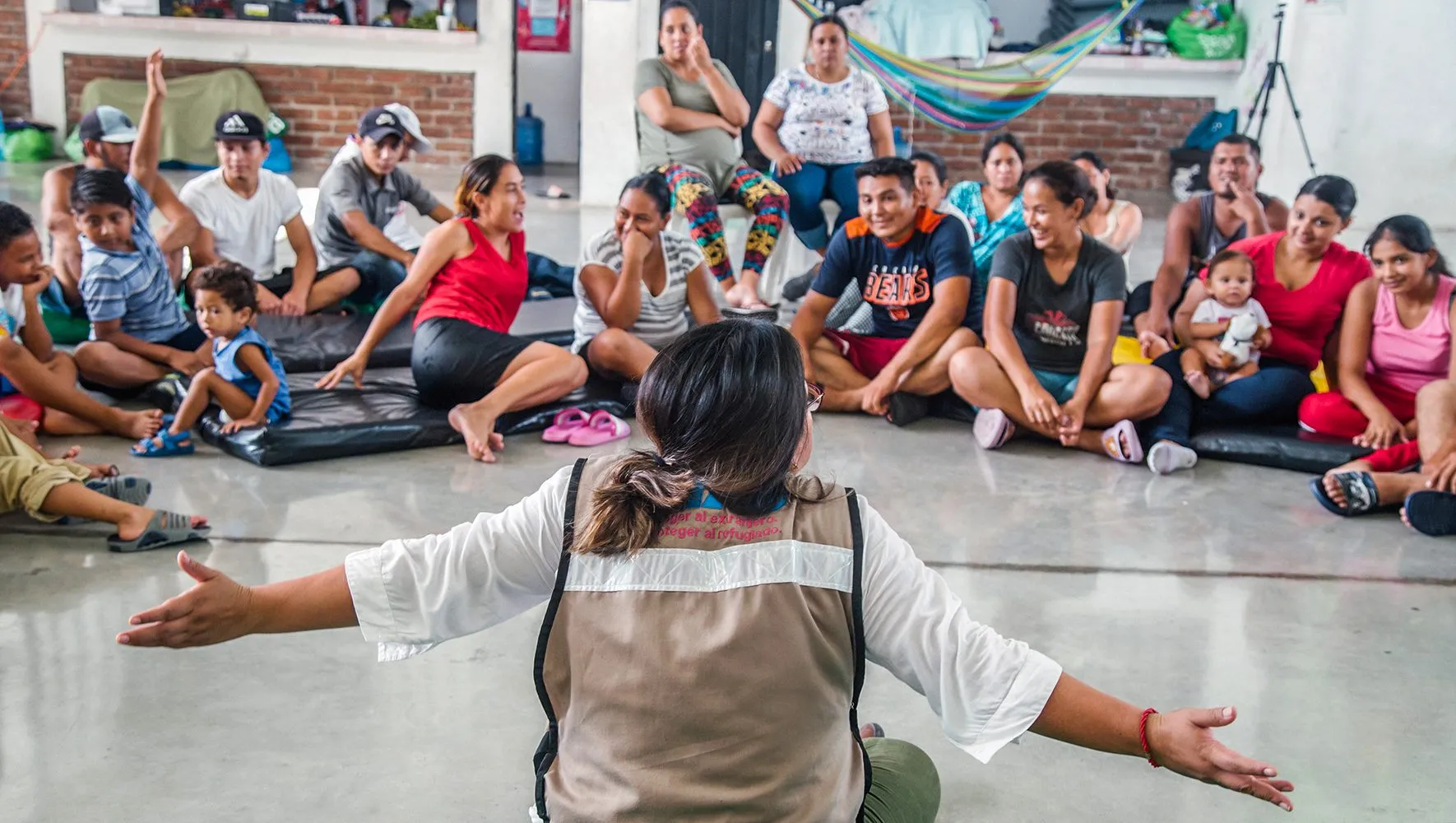 Një grup njerëzish të ulur në dysheme në një mjedis komunitar, dëgjojnë me vëmendje një profesioniste që flet me krahë të hapur, duke treguar mbështetje dhe komunikim të hapur.