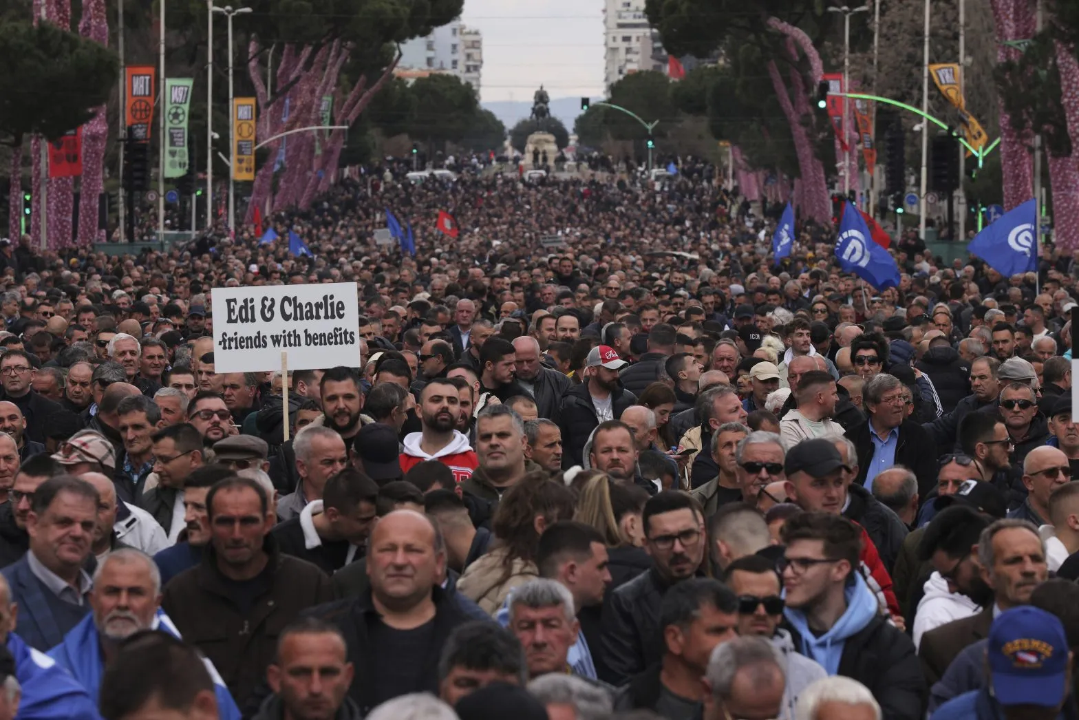 Një turmë e madhe protestuesish opozitarë marshon në rrugët e Tiranës gjatë një demonstrimi kundër qeverisë, me një tabelë që lexon 'Edi & Charlie friends with benefits' e mbajtur në plan të parë.