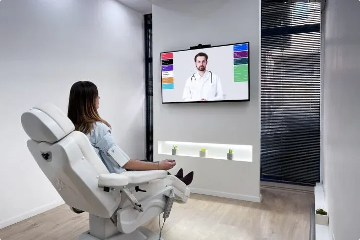Një person ulur në një klinikë dixhitale duke komunikuar me një mjek në një ekran të madh, duke shfaqur modernizimin e shërbimeve.
