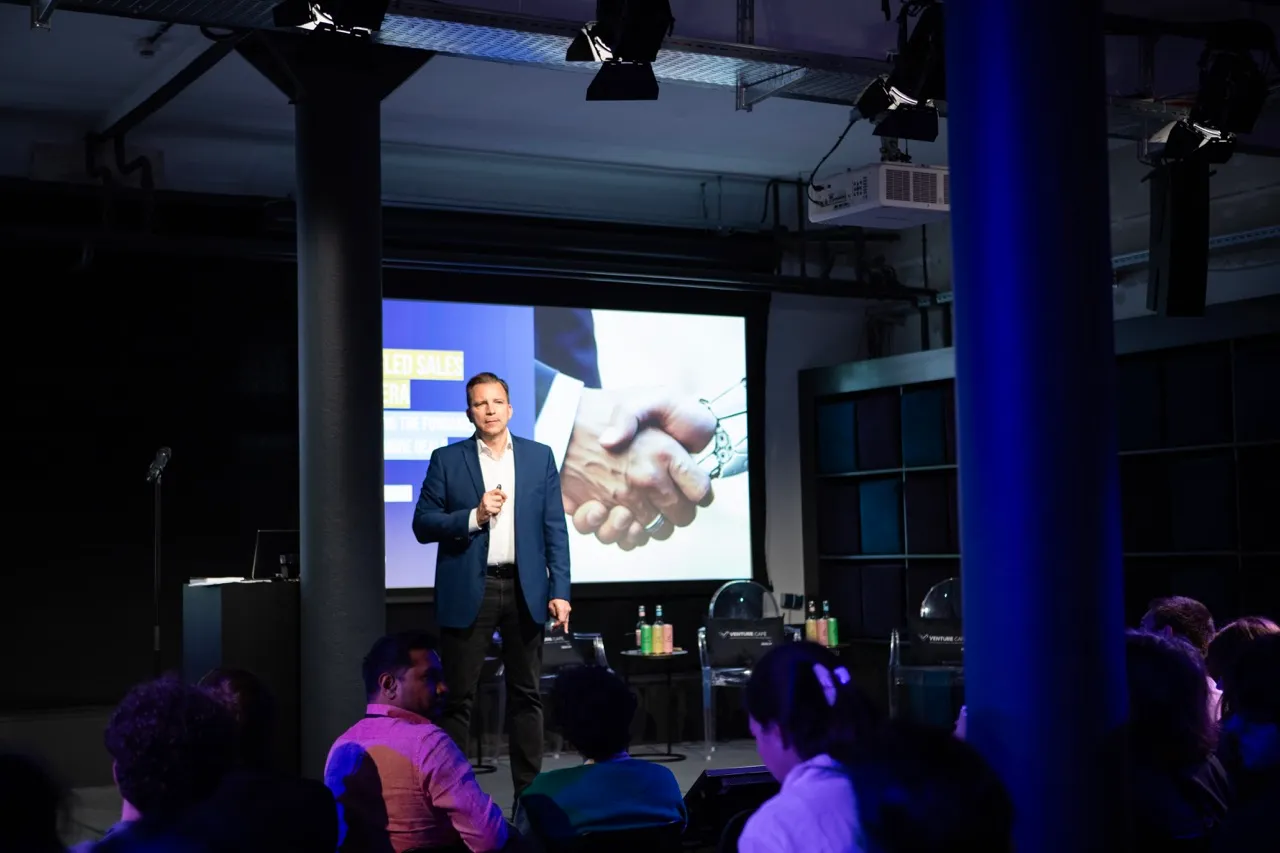 Një folës në një konferencë teknologjike, duke prezantuar para një audience, me një imazh digjital bashkëpunimi në sfond.