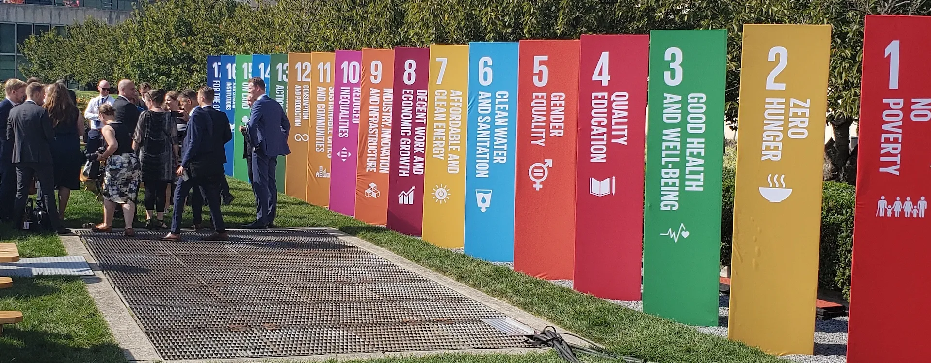 Njerëz që qëndrojnë pranë shtyllave shumëngjyrëshe që përfaqësojnë Qëllimet e Zhvillimit të Qëndrueshëm të OKB-së, duke simbolizuar zhvillimin ekonomik dhe social afatgjatë.