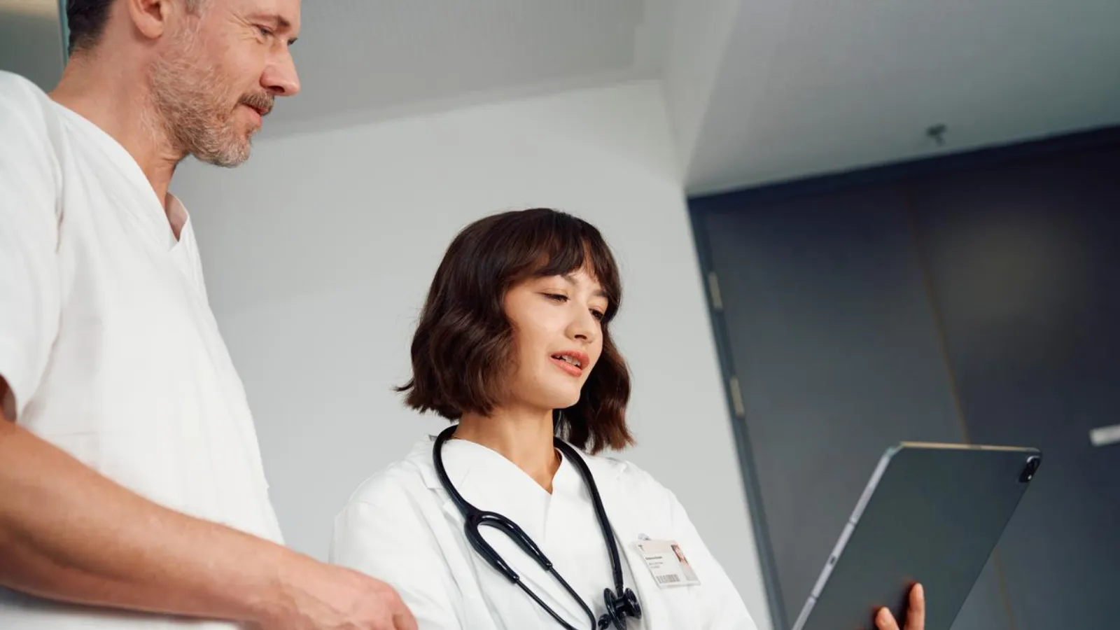 Dy profesionistë shëndetësorë, një burrë dhe një grua, duke parë një tablet së bashku në një ambient spitalor, duke simbolizuar kujdesin mjekësor modern.