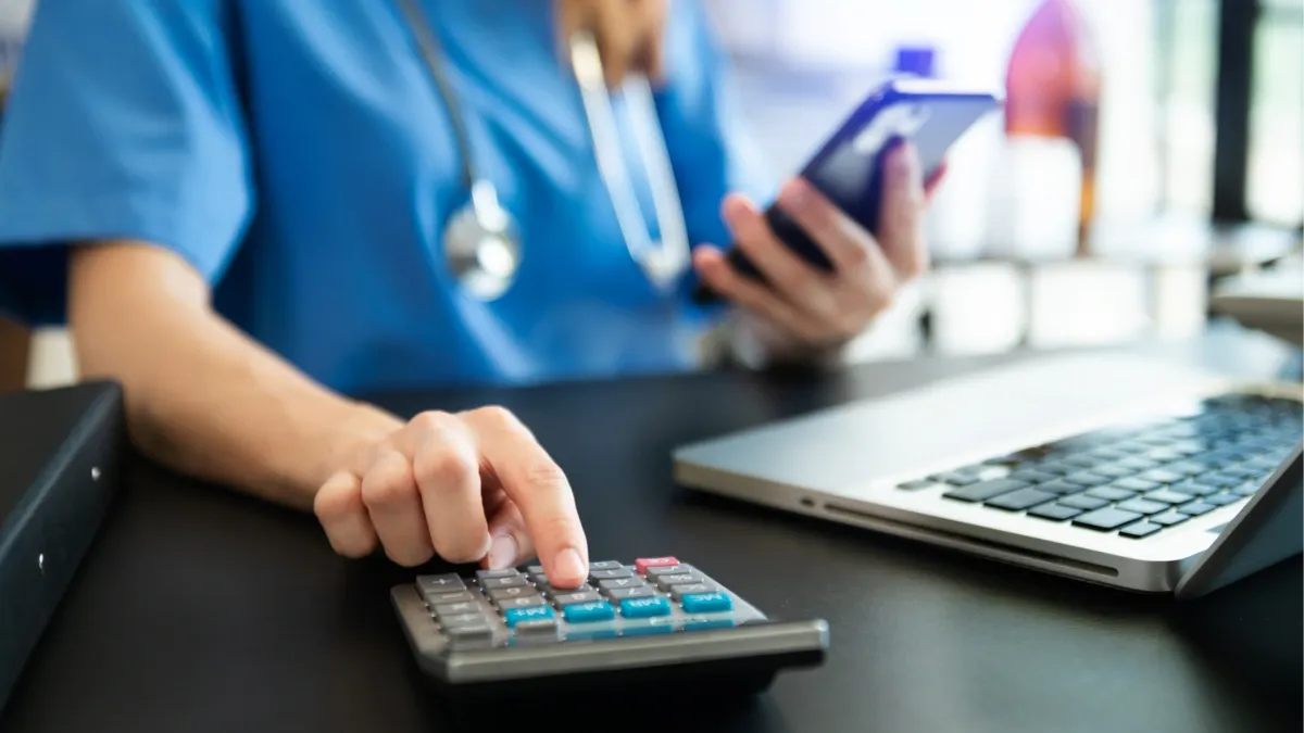 Duart e një personi duke përdorur një kalkulator dhe laptop, duke simbolizuar shpenzimet dhe kostot e kujdesit shëndetësor.