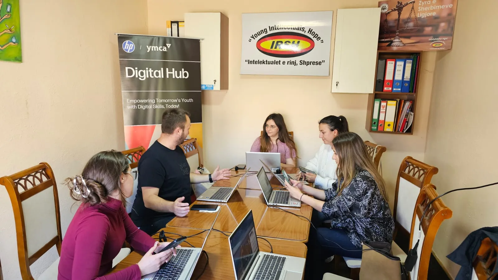 Të rinj duke punuar me pajisje dixhitale në një hapësirë bashkëpunuese, me mbishkrimin 'Digital Hub' në sfond, duke theksuar aftësimin dixhital.