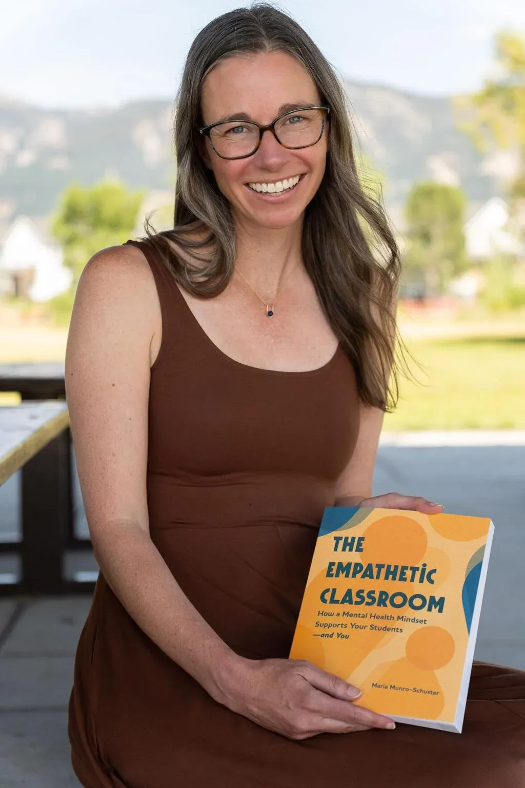 Një grua e buzëqeshur mban një libër me titull "Klasa Empatike", duke theksuar rëndësinë e edukimit dhe ndërgjegjësimit për shëndetin mendor.